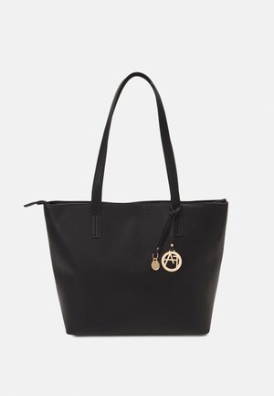 Sac cabas noir avec deux longues anses et un charm circulaire doré suspendu à une anse sur un fond uni.
