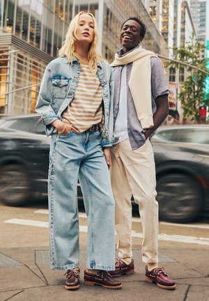 Ein Paar Models steht im Freien. Eines trägt eine leichte Jeansjacke, ein gestreiftes Hemd und weit geschnittene Jeans; das andere trägt ein graues Hemd und cremefarbene Hosen. Braune Schuhe.