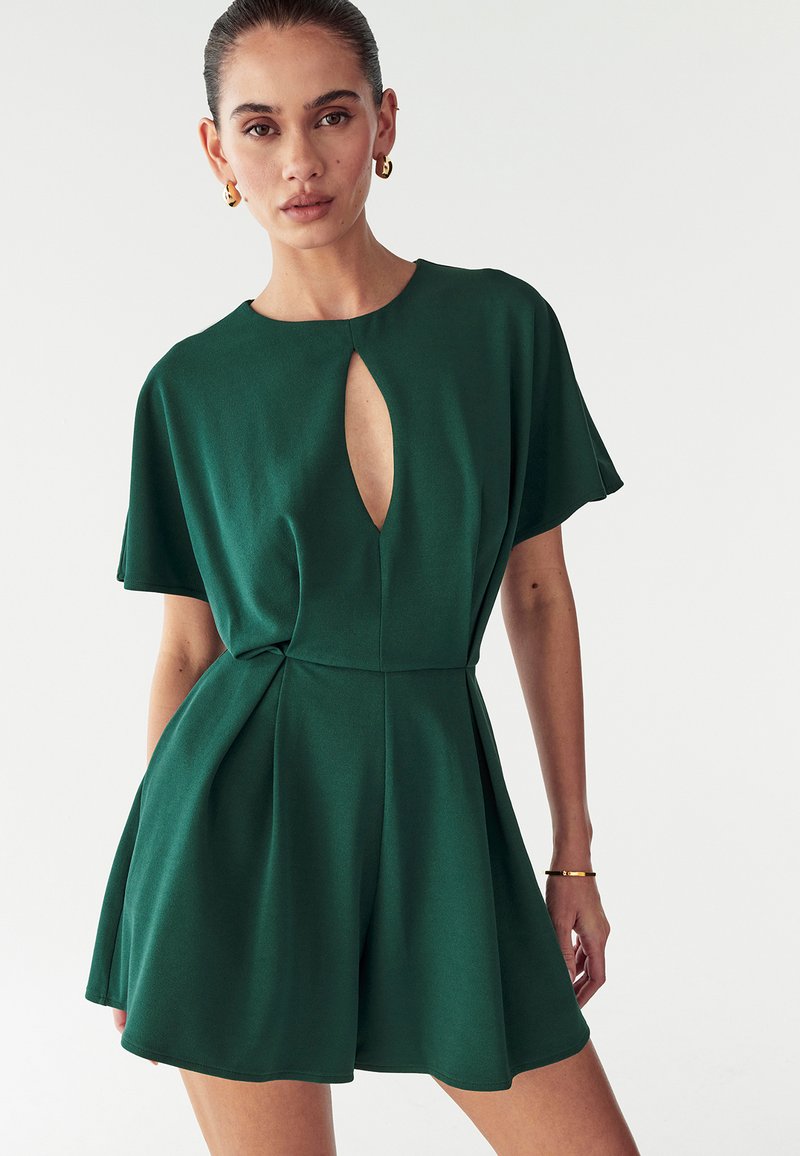 Robe courte verte avec un décolleté à découpe, manches courtes et jupe plissée. Fabriquée en tissu lisse, elle met en valeur une taille cintrée.