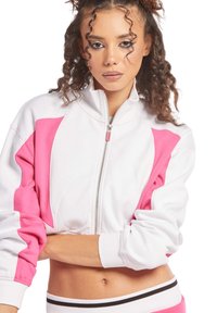 Giacca a zip cropped bianca con dettagli rosa e polsini a coste. Presenta un colletto alto e una texture liscia. Il modello posa con le braccia incrociate.
