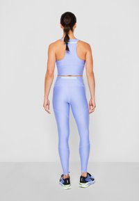 Žena stojící zády, oblečená v světle fialovém sportovním podprsence a legínách s černými a fialovými běžeckými botami na jednobarevném pozadí.