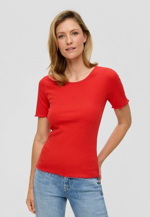 Femme blonde portant un haut rouge ajusté à manches courtes avec des bords volantés et un jean bleu clair, se tenant devant un fond gris uni.