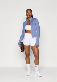 Giacca sportiva azzurro chiaro a maniche lunghe, top corto bianco e pantaloncini bianchi. Bottiglia d'acqua nera tenuta in una mano, sneakers bianche abbinate.