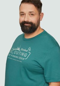 Turkusowa bawełniana koszulka z okrągłym dekoltem, krótkimi rękawami oraz białym nadrukiem graficznym z motywem wędkarskim i elementami tekstowymi.