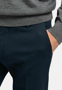 Mörkblå texturerade byxor med sidofickor, matchade med en ljusgrå tröja med ribbat avslut. Närbild av midjan och handen i fickan.