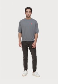 Giovane uomo in piedi, che indossa una maglietta grigia a maniche lunghe con un piccolo logo arancione, pantaloni neri e sneakers bianche su uno sfondo bianco semplice.