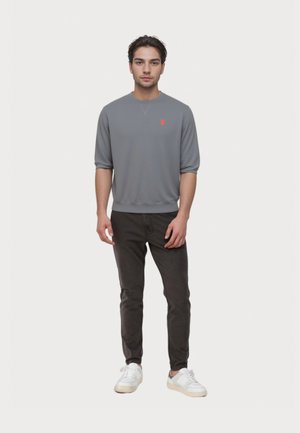 Jonge man staat, draagt een grijs long sleeve shirt met een klein oranje logo, zwarte broek en witte sneakers op een effen witte achtergrond.