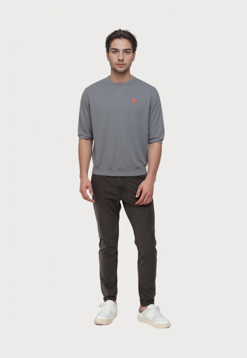Młody mężczyzna stoi, ubrany w szary t-shirt z długim rękawem z małym pomarańczowym logo, czarne spodnie i białe sneakersy, na jednolitym białym tle.