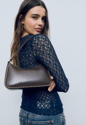 Una giovane donna indossa un top di pizzo blu navy e jeans, mentre tiene sulla spalla una borsa in pelle marrone scuro con borchie metalliche.