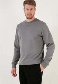 Felpa grigia con collo rotondo e polsini a costine. Presenta una sottile texture e un dettaglio di cucitura a triangolo sul collo. Abbinata a pantaloni neri.