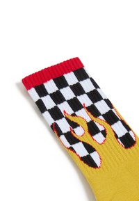 Żółta skarpetka z czerwono-czarną szachownicą u góry, z grafiką czerwonych płomieni w okolicy kostki. Miękka, dziana tekstura.