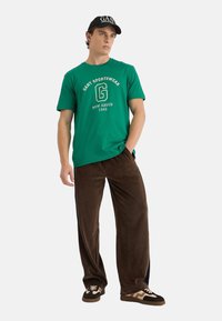 Camiseta de algodón verde con logo blanco, combinada con pantalones de terciopelo marrón. Los pantalones tienen un corte holgado y slit laterales. Zapatillas blancas y negras.