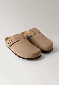 Beige suede clogs with a rounded toe, a single adjustable buckle strap, and a textured rubber sole. Smooth finish with a minimal design.