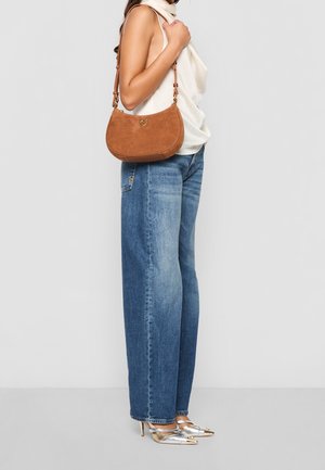 Femme portant un jean bleu, un haut blanc sans manches, des talons hauts argentés, et portant un sac à épaule en daim marron avec une fermeture dorée.