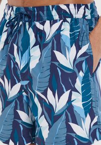 Costume da bagno con un motivo floreale blu e grandi foglie tropicali bianche. Realizzato in tessuto leggero con una vita elastica e tasche.