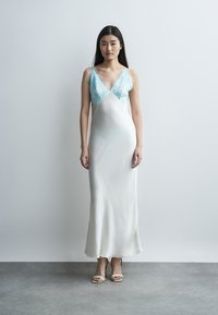 Νεαρή γυναίκα στέκεται σε λευκό τοίχο, φορώντας μακρύ σατέν φόρεμα με μπλε δαντελένιο μπούστο και μπεζ πέδιλα με τακούνι.