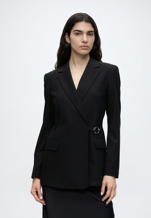 Femme aux longs cheveux foncés portant un blazer noir ajusté avec un détail d’anneau sur le côté, debout devant un fond clair uni.