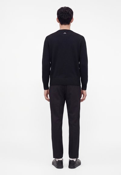 Homme debout de dos portant une chemise noire à manches longues, un pantalon noir, des chaussettes blanches et des chaussures noires sur fond blanc.