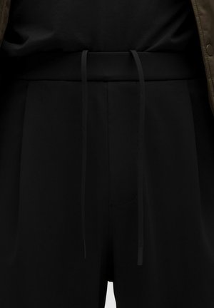 Pantalones negros con una cinturilla de tela y cordones largos, llevados con una parte superior negra y una chaqueta marrón.