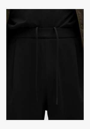 Pantalones negros con una cinturilla de tela y cordones largos, llevados con una parte superior negra y una chaqueta marrón.