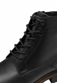 Bottine en cuir noir avec des lacets ronds noués en nœud, œillets en métal et couture visible sur le côté et la languette.