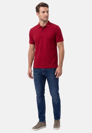 Uomo con polo a maniche corte rossa, jeans blu e sneaker taupe, in piedi con le mani rilassate, guarda verso destra.