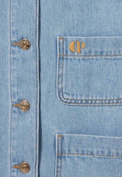 Un tissu en denim bleu clair présente des boutons-pression dorés et une poche plaquée. Des surpiqûres orange mettent en valeur les bords de la poche et les détails du logo.