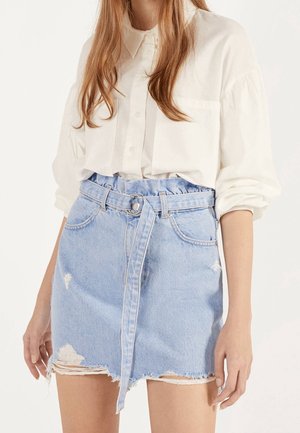 Vrouw die een wit overhemd met knopen draagt met opgerolde mouwen, ingestopt in een lichtblauwe versleten denim rok met een bijpassende riem.