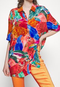 Kvinna som bär en färgglad skjorta med tryck av tropiska djur och vegetation, i kombination med klara orange byxor.
