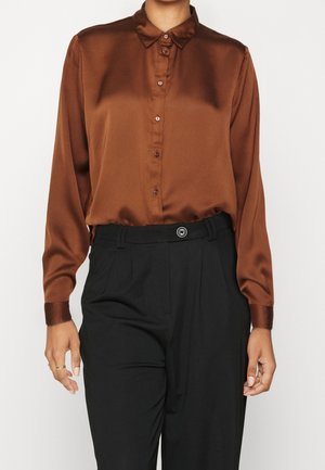 Femme portant une chemise en satin marron à manches longues boutonnée, rentrée dans un pantalon noir taille haute avec une fermeture à un seul bouton.