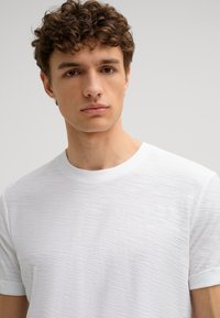 Weißes Baumwoll-T-Shirt mit strukturierter Oberfläche, ribbbeltem Kragen und umgeschlagenen kurzen Ärmeln, das einen lässigen Schnitt und ein minimalistisches Design aufweist.