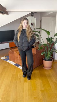 Melna pufjakas jaka ar rāvējslēdzēju un sānu kabatām, kopā ar melnām, plašām biksēm un melniem apaviem. Redzams koka grīdas segums un istabas augi.