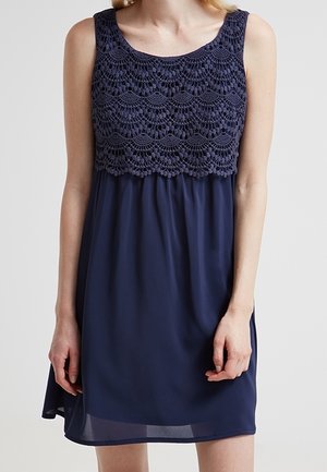 Kobieta ubrana w granatową sukienkę bez rękawów z koronkowym gorsetem na szydełku i zwiewną spódnicą z szyfonu, stojąca na jednolitym tle.