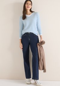 Femme debout contre un mur beige, portant un pull bleu clair, un jean foncé, des baskets blanches, et tenant une veste marron dans sa main droite.