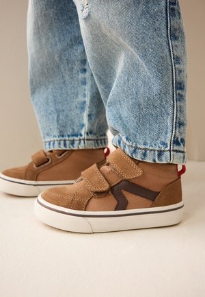 Kind trägt braune Klett-Sneaker mit weißen Sohlen und blaue Jeans und steht auf einem hellen Boden.