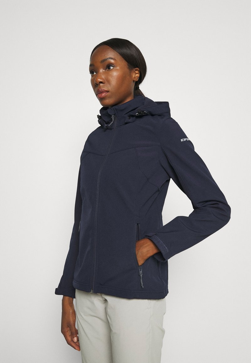 Veste softshell bleu marine avec poches zippées, capuche et cordons de serrage ajustables. Elle présente une texture lisse et une coupe athlétique.