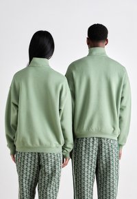 Two people stand back facing, wearing sage green high-neck sweatshirts and patterned green and white track pants.