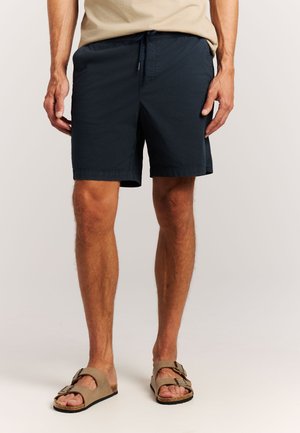 Man die beige sandalen en zwarte knielange korte broek draagt, met licht gespreide benen staand tegen een effen achtergrond.