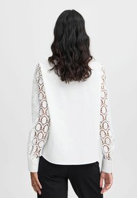 White blouse with sheer lace sleeves, featuring intricate circular patterns. The body is solid white, and it has fitted cuffs.