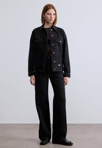 Μαύρο τζιν μπουφάν με μπροστινές τσέπες και μεταλλικά κουμπιά, σε συνδυασμό με μαύρο τζιν παντελόνι ίσιας γραμμής και μαύρα παπούτσια, τοποθετημένα σε ουδέτερο φόντο.