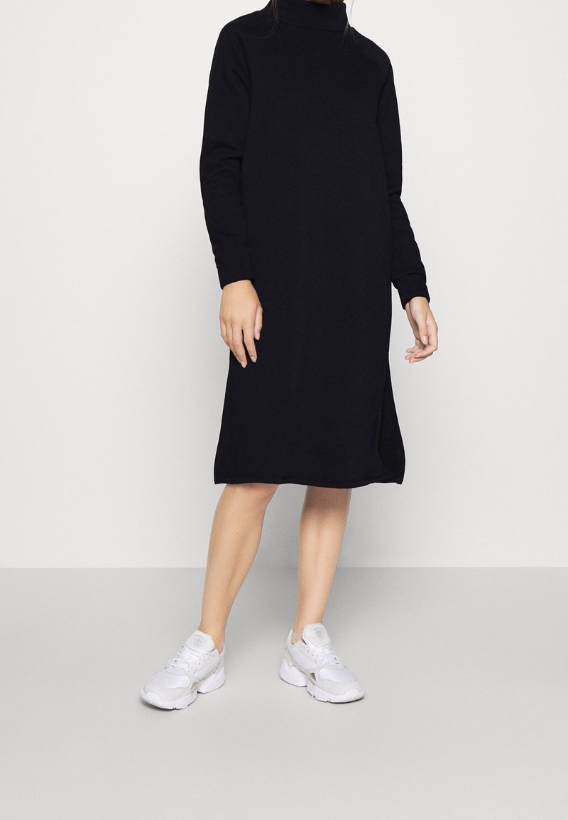 Personne portant une robe noire à manches longues, longueur genou, avec un col montant et des baskets blanches, debout devant un fond uni.