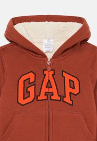 Brun zip-up hette med kremfarget fleecefôr, store oransje og svarte "GAP" bokstaver foran, og to frontlommer.
