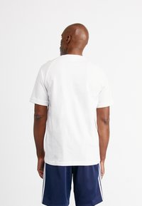 Valkoinen puuvillainen T-paita, jossa on lyhyet hihat ja pyöreä pääntie, nähtynä takaa. Yhdistetty tummansinisiin shortseihin, joissa on valkoiset sivuraidat.