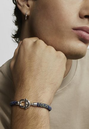 Pulsera de cierre de plata con tela trenzada en patrones azul, amarillo y verde. Primer plano de una mano descansando sobre la mejilla, usando un pendiente de aro de plata.