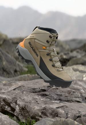 Világosbarna és fekete Scarpa túracipő narancssárga díszítésekkel, Gore-Tex címkével, masszív talppal, sziklás terep fölött elhelyezve, háttérben elmosódott hegyek.