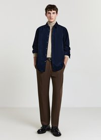 Camicia in velluto a coste blu navy indossata sopra un dolcevita beige, abbinata a pantaloni marroni e scarpe nere, creando un outfit testurizzato e a contrasto.