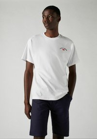 Vit t-shirt i bomull med röd och svart text, kombinerad med mörkblå shorts. Enkelt design, rund halsringning, korta ärmar och avslappnad passform.