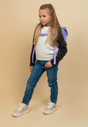 Weißer gestrickter Pullover mit schwarzen Ärmeln, blaue Jeans und weiße Sneaker. Lila Rucksack, geflochtenes Haar, Stehpose vor einem beigen Hintergrund.