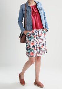 Frau in einer Jean-Jacke, rotem Blusenshirt, Blumenkleid, braunen Espadrilles und mit einer braunen Schultertasche, die mit Händen in den Taschen steht.