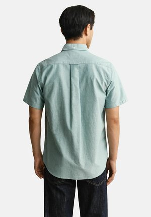 Mann, der ein hellgrünes kurzärmeliges Hemd mit Knöpfen und dunkle Jeans trägt, von hinten vor einem weißen Hintergrund zu sehen.